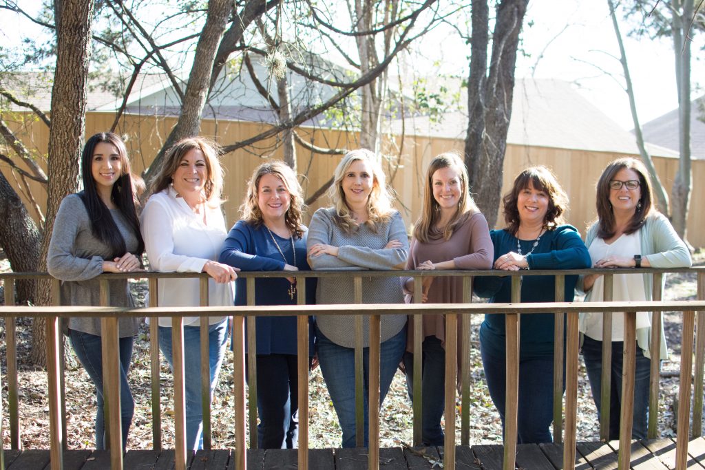 The Smartt Team is lined up posing outside for a team photo. Pictured from left to right is Adel Gould, Patti McDaniel, Paula Durocher, Amy Nelson, Marla McDonald, Irene Norton, and Pam Smartt. Behind the team is a fence, tall pine trees, and leaves fallen on the ground.