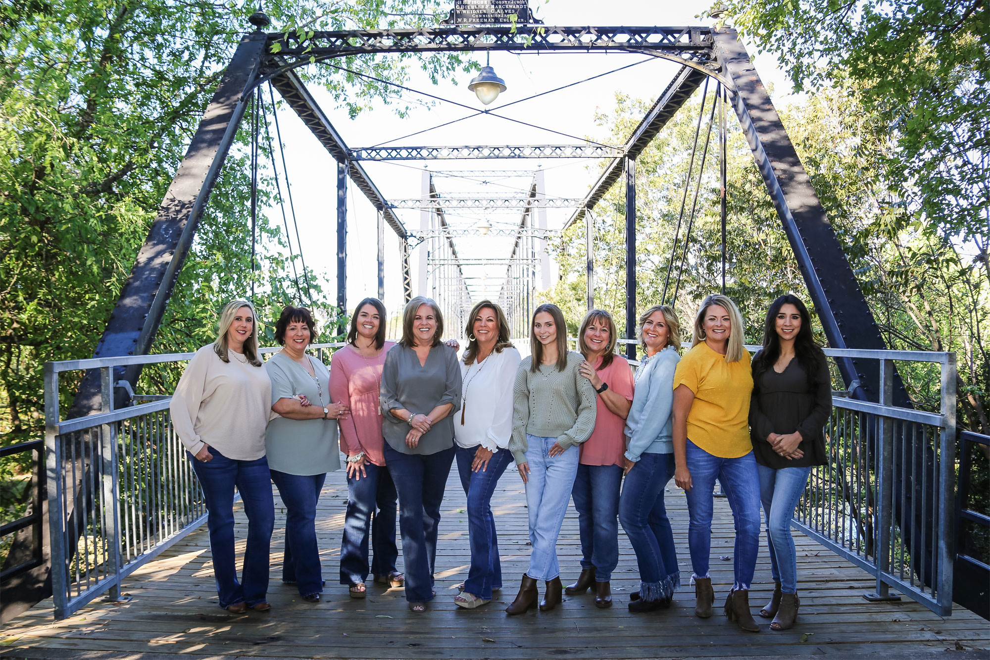 The Smartt Team is lined up posing outside for a team photo. Pictured from left to right is Adel Gould, Patti McDaniel, Paula Durocher, Amy Nelson, Marla McDonald, Irene Norton, and Pam Smartt. Behind the team is a fence, tall pine trees, and leaves fallen on the ground.